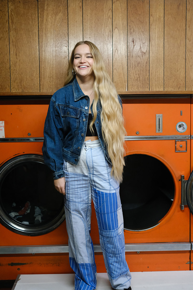a whole body picture of Charlotte Jane in front of old, orange washing machines wearing a jeans look. It's the same location as her 2020 EP-cover.